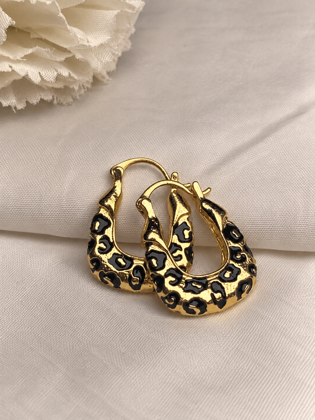 Gold-tone chunky hoop earrings with glossy black abstract enamel patterns, featuring a bold sculptural design on soft cream fabric.