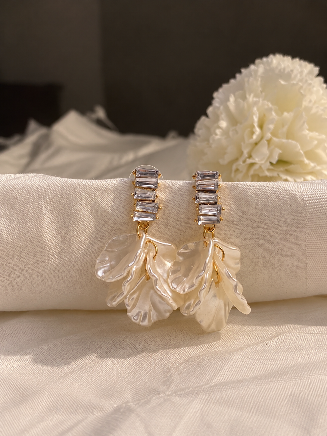 Gold-tone dangling earrings with stacked clear crystal accents and translucent white floral petal drops, displayed on soft cream fabric.