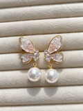 Gold-tone butterfly earrings with sparkling clear and blush-pink crystals, finished with large white pearl drops, displayed on soft cream fabric.