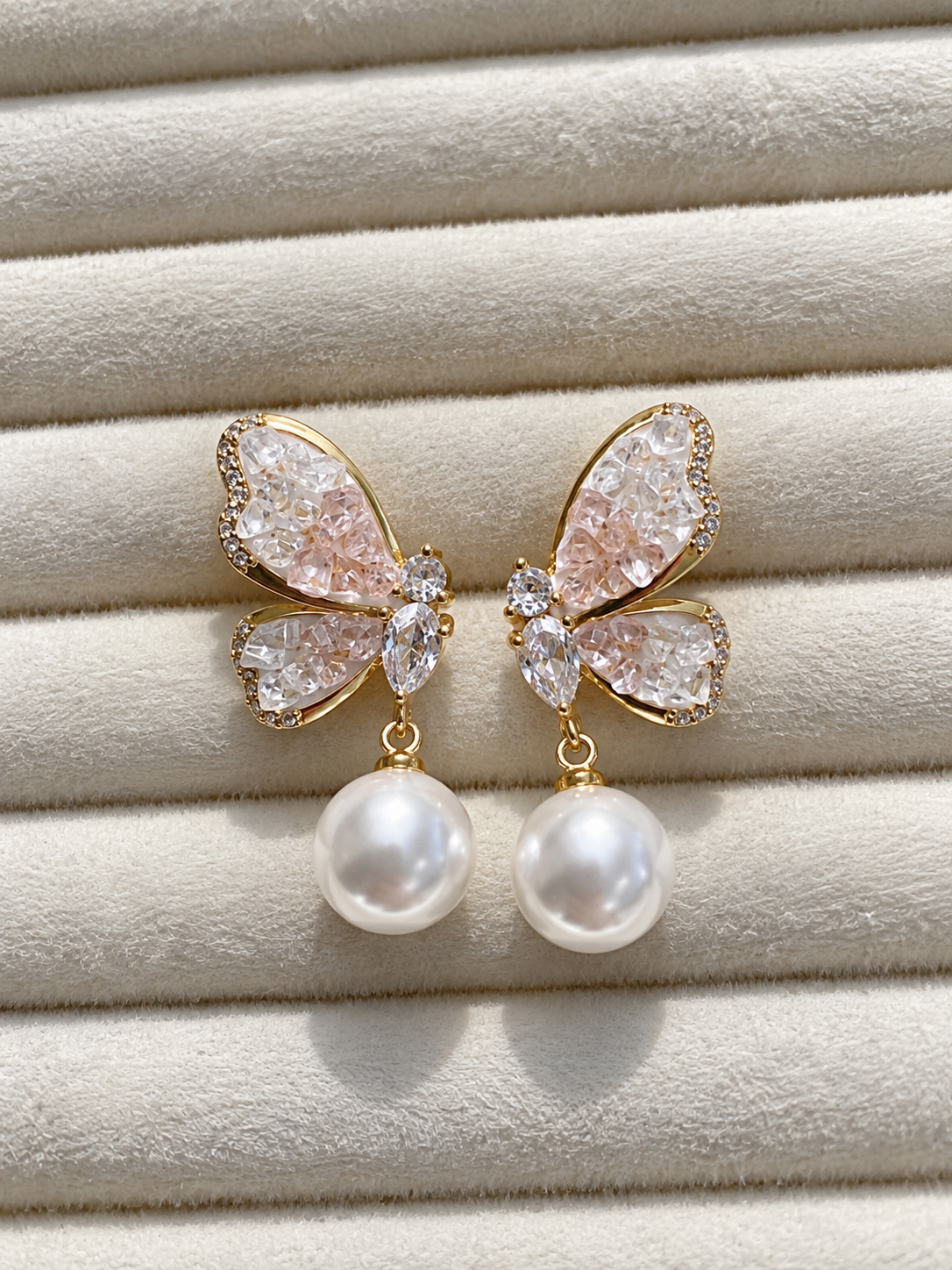 Gold-tone butterfly earrings with sparkling clear and blush-pink crystals, finished with large white pearl drops, displayed on soft cream fabric.