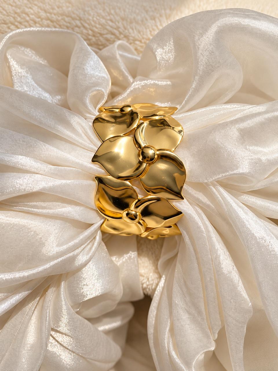 A polished gold flower-shaped hair claw clip displayed on a voluminous cream satin scrunchie.