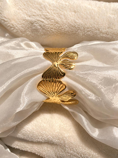 A gold-toned metal cuff bracelet with three textured, fan-like leaf or ginkgo-inspired segments, displayed on draped white satin fabric over a soft cream fleece background.