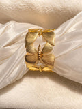 A gold butterfly-shaped cuff bracelet with textured, ribbed wings, displayed on a cream-colored silk ribbon over a soft beige fabric background.
