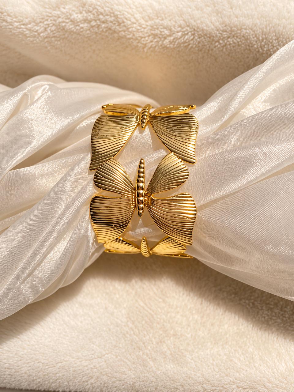 A gold butterfly-shaped cuff bracelet with textured, ribbed wings, displayed on a cream-colored silk ribbon over a soft beige fabric background.