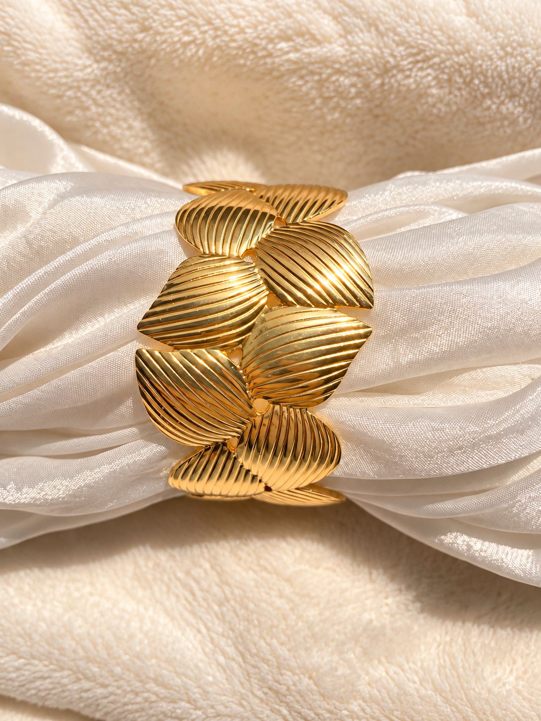 A wide gold cuff bracelet with a textured, ribbed leaf-like design, displayed on draped white satin fabric.