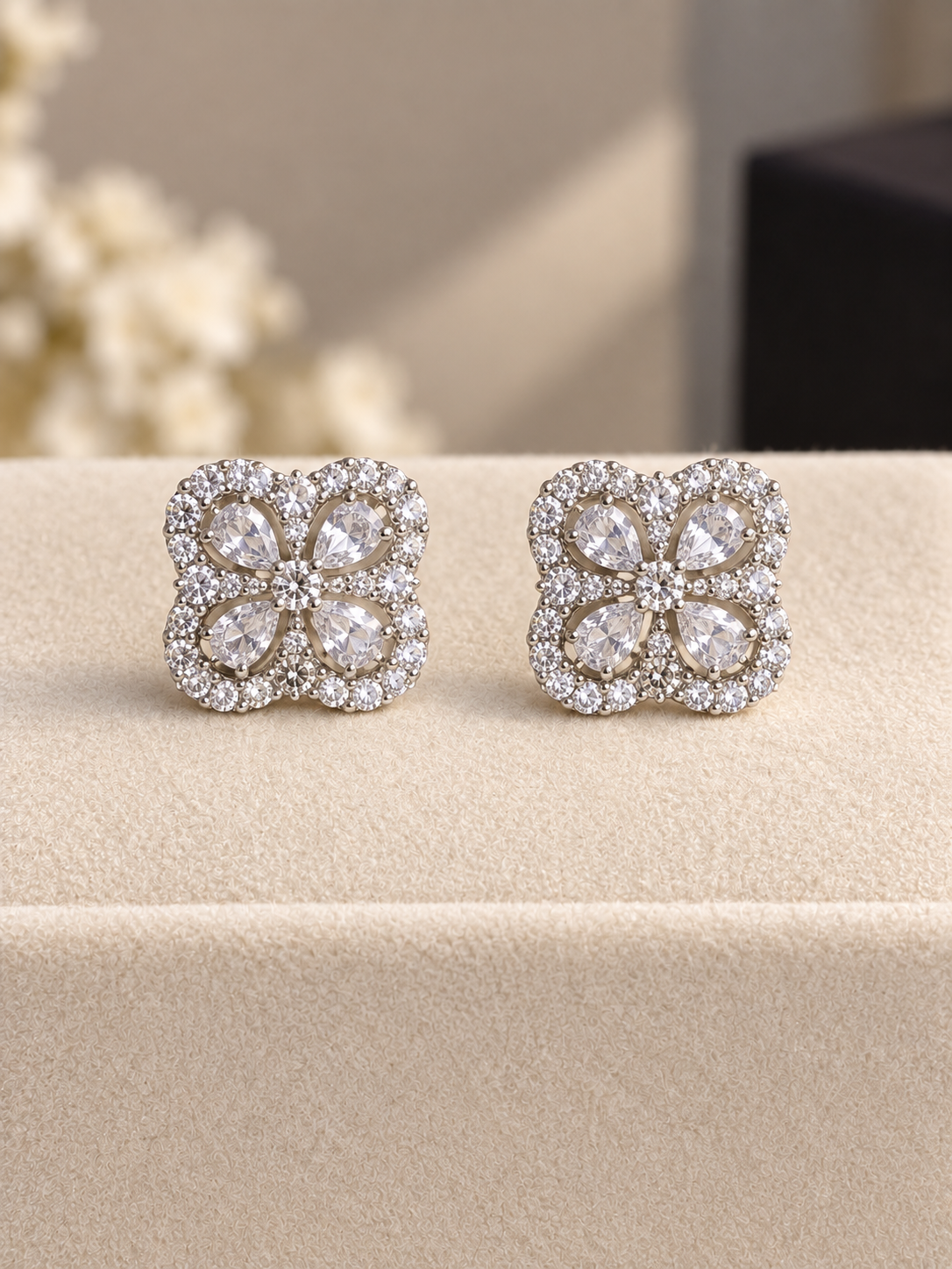 Silver-tone square stud earrings featuring four-petal floral designs with sparkling clear teardrop-shaped crystals and smaller crystal accents, displayed on soft cream fabric.