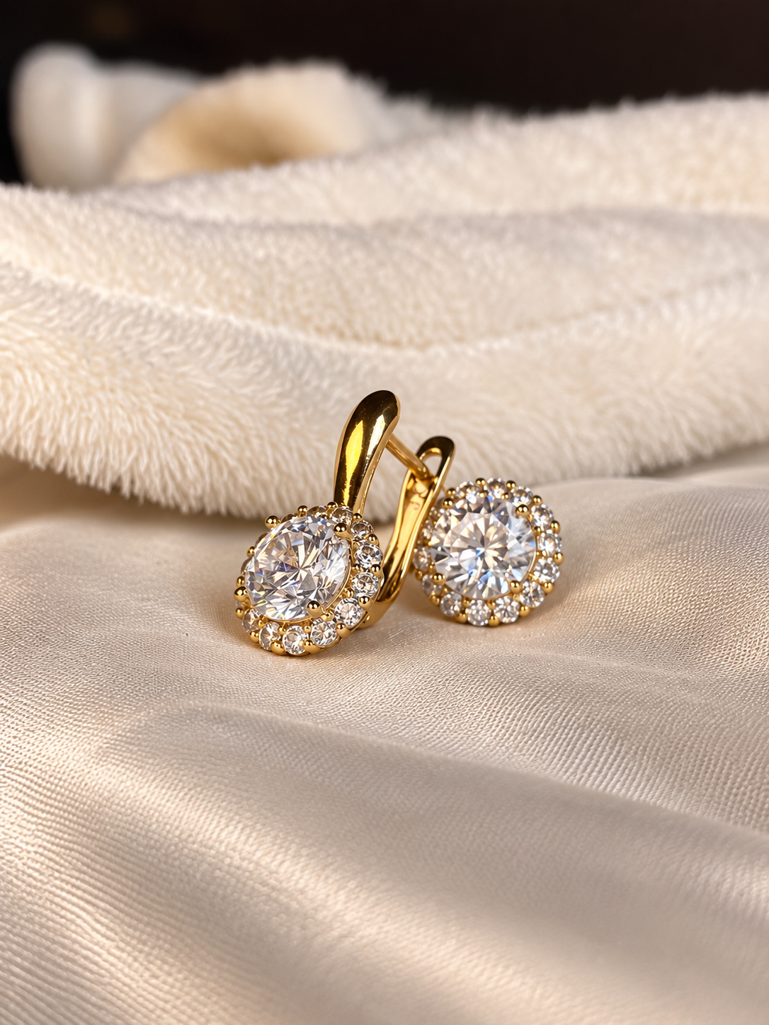 A pair of gold hoop earrings with oval halo settings set with clear gemstones, resting on a cream satin fabric with a soft plush cream background.