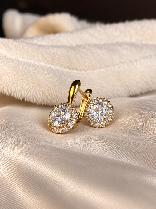 A pair of gold hoop earrings with oval halo settings set with clear gemstones, resting on a cream satin fabric with a soft plush cream background.