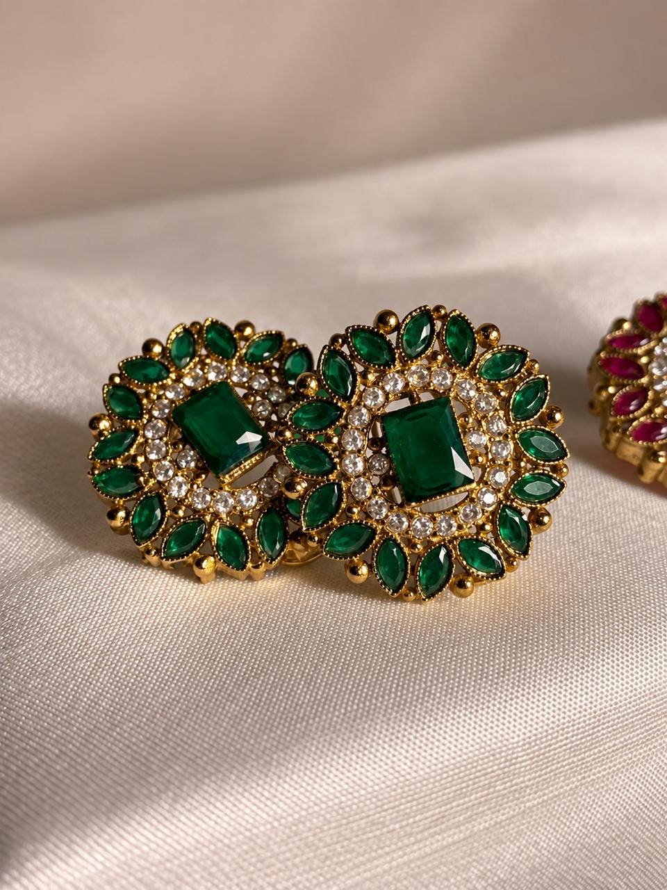 A pair of ornate gold-toned statement earrings featuring green emerald-cut center stones surrounded by green marquise-shaped stones and small clear crystals, displayed on a beige satin fabric.