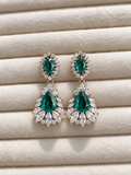 merald green drop earrings with large faceted teardrop stones, surrounded by sparkling clear crystals and silver-tone detailing, displayed on soft cream fabric.