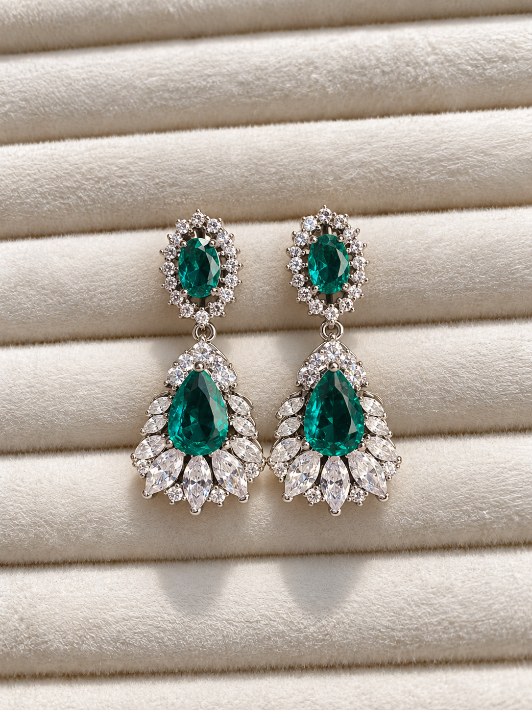 merald green drop earrings with large faceted teardrop stones, surrounded by sparkling clear crystals and silver-tone detailing, displayed on soft cream fabric.