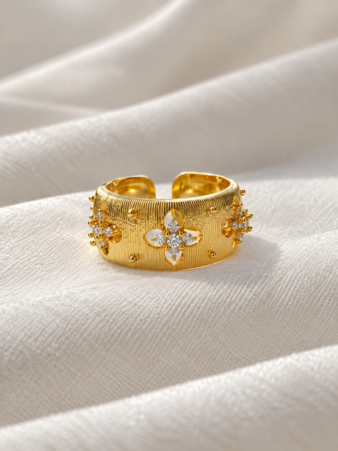 Adjustable gold-tone statement ring featuring textured floral motifs with sparkling clear crystals, displayed on soft white fabric.