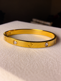 Gold-tone bangle bracelet featuring alternating clear crystal accents and embossed four-petal floral motifs, displayed on soft beige fabric.