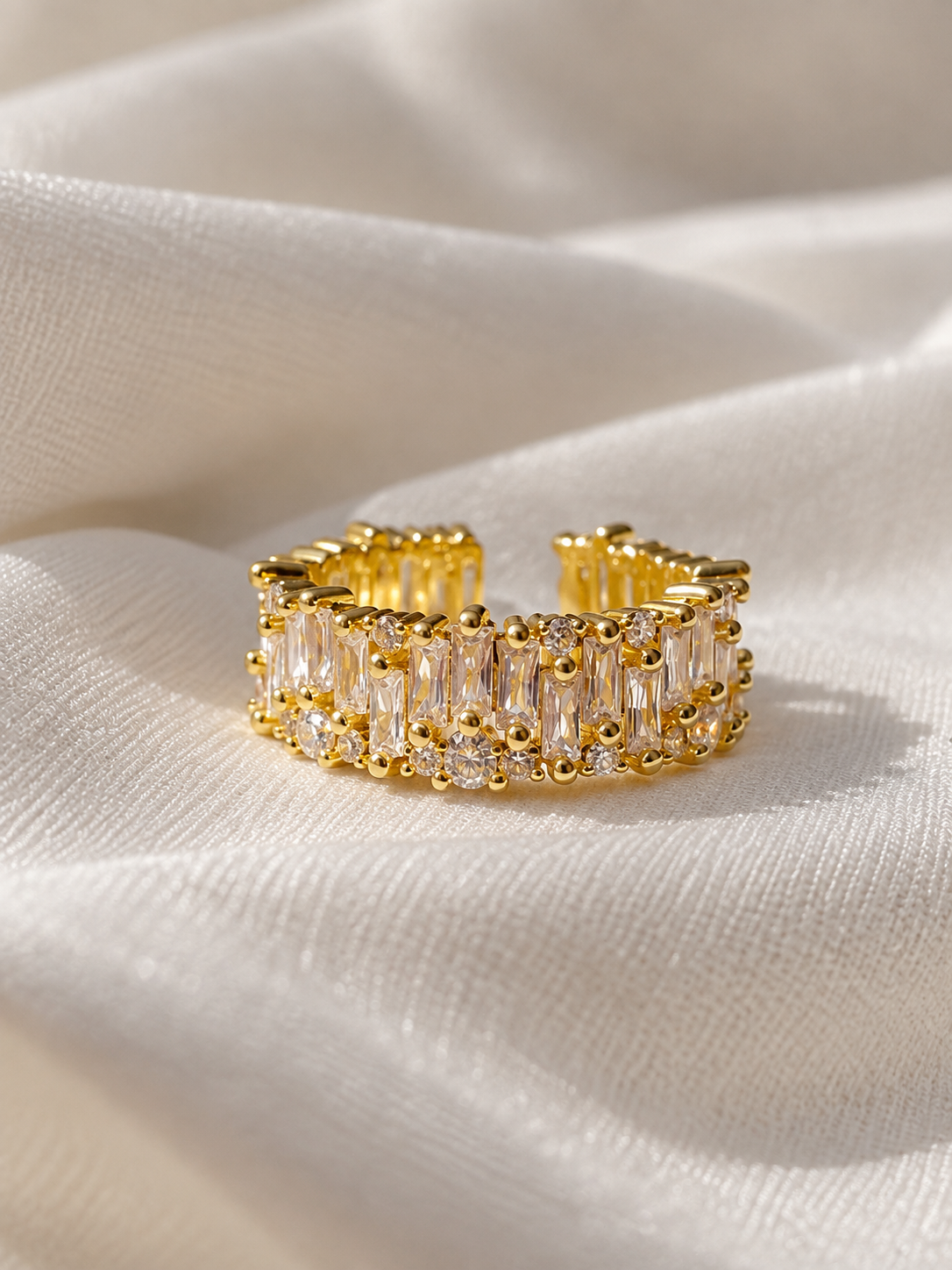 Adjustable gold-tone ring featuring rows of sparkling clear baguette-cut crystals and smaller round crystals, displayed on soft cream fabric.