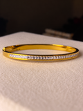 Slim gold-tone cuff bracelet featuring a continuous row of clear baguette-cut crystals along the front, displayed on soft beige fabric.