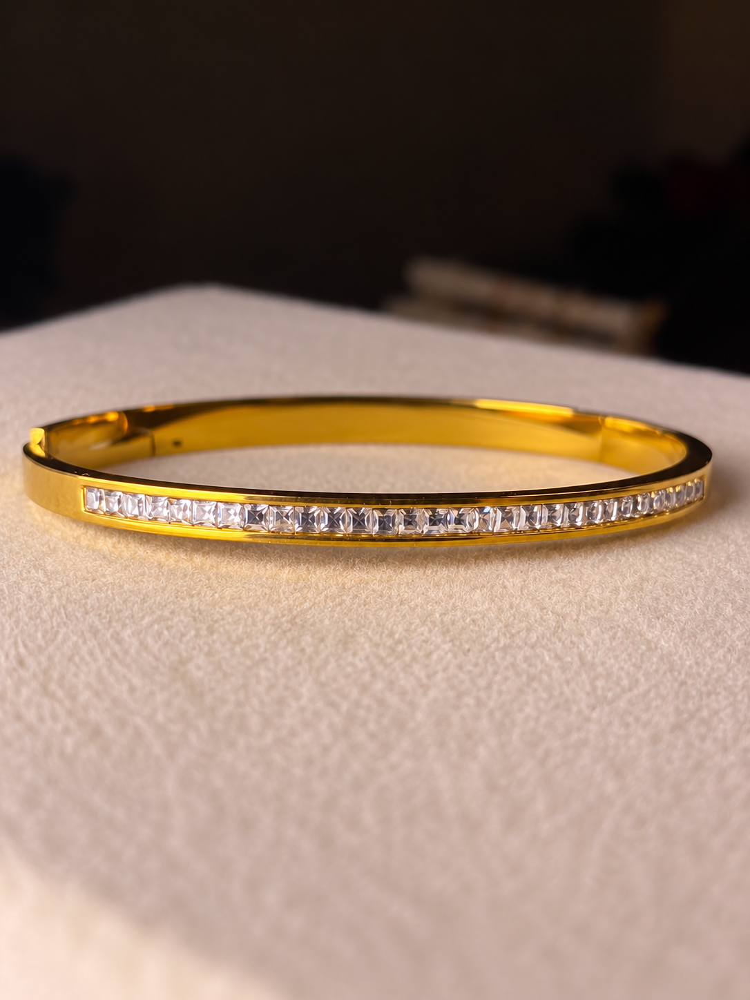 Slim gold-tone cuff bracelet featuring a continuous row of clear baguette-cut crystals along the front, displayed on soft beige fabric.