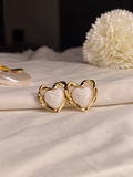 A pair of gold heart-shaped stud earrings with white textured inlay, displayed on a draped cream fabric next to a white flower.