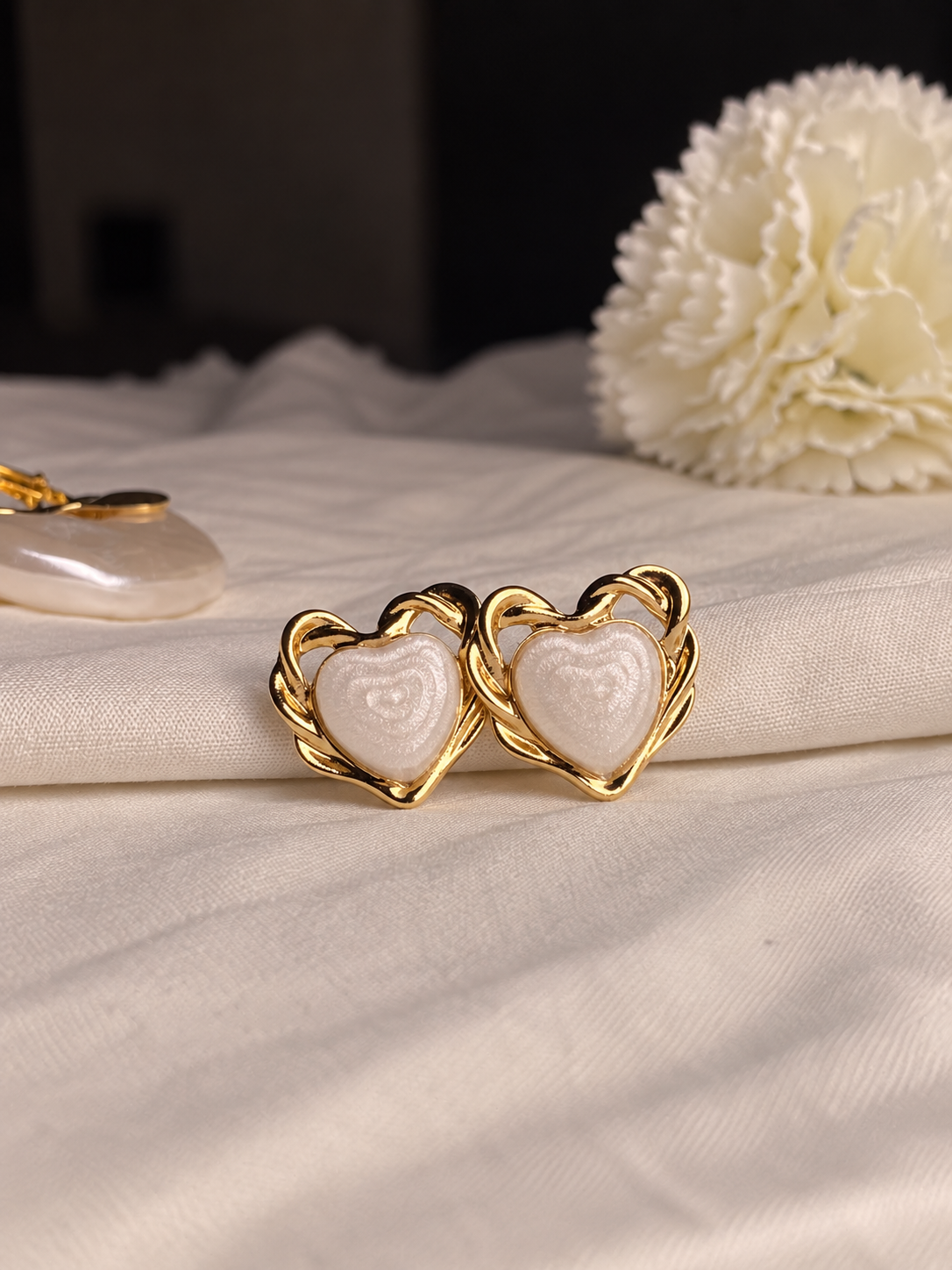 A pair of gold heart-shaped stud earrings with white textured inlay, displayed on a draped cream fabric next to a white flower.