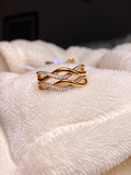 Gold-tone statement ring with intertwined curved bands, featuring a row of sparkling clear crystals and a polished finish, displayed on soft white fabric.