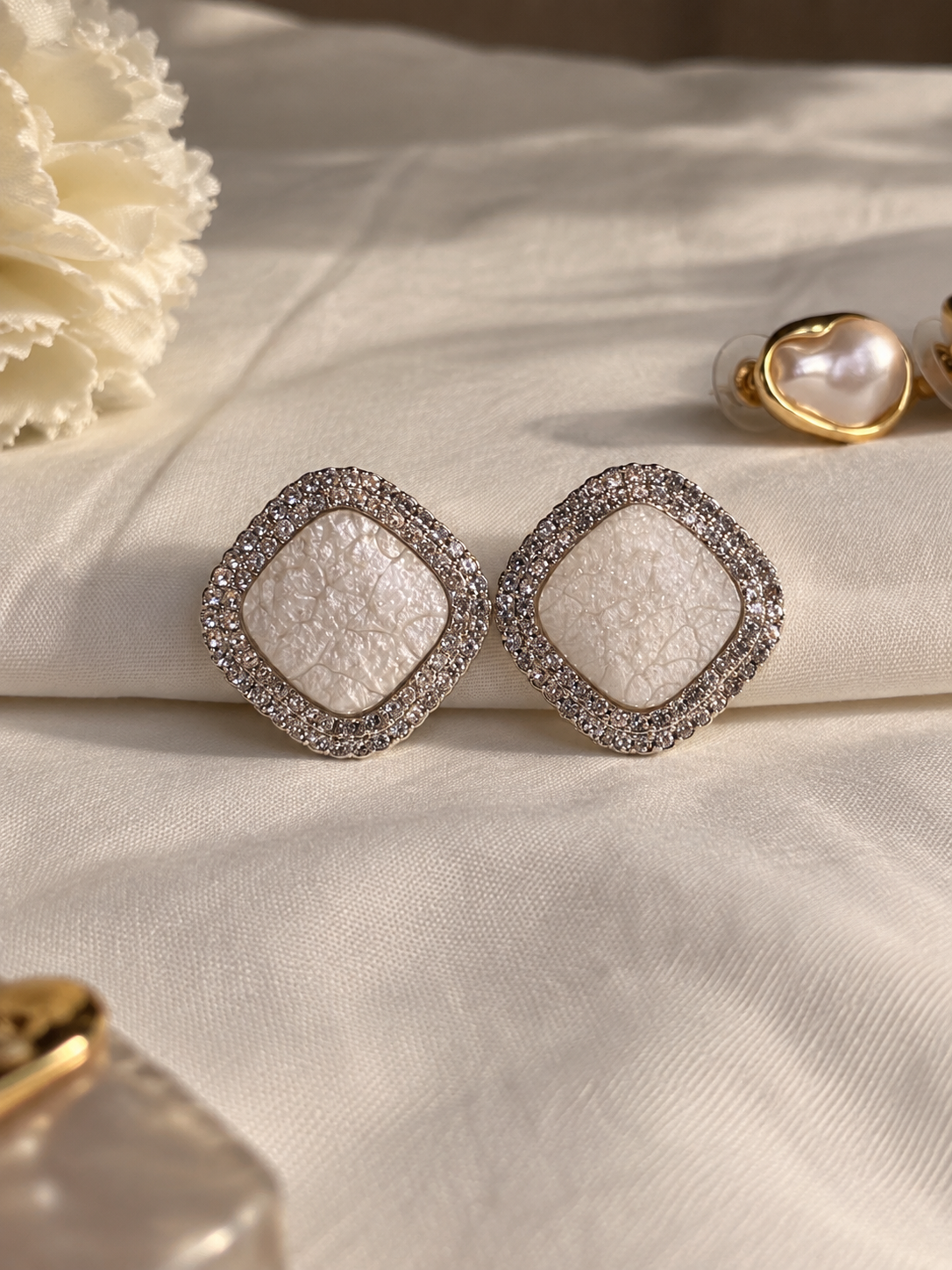 Ivory square stud earrings with a textured pearl-like center, surrounded by sparkling clear crystals and a polished silver-tone border.