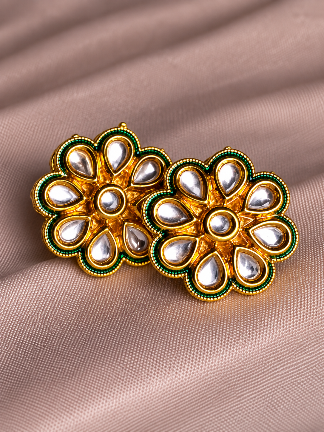 A pair of gold-toned floral Kundan earrings with silver teardrop stones and green border accents, displayed on a draped beige fabric background.