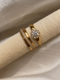 Three gold rings displayed on a textured cream fabric ring holder. One ring features a round brilliant-cut diamond in a halo setting, while the other two are slim bands, one with a pavé band and one with a curved row of small diamonds.