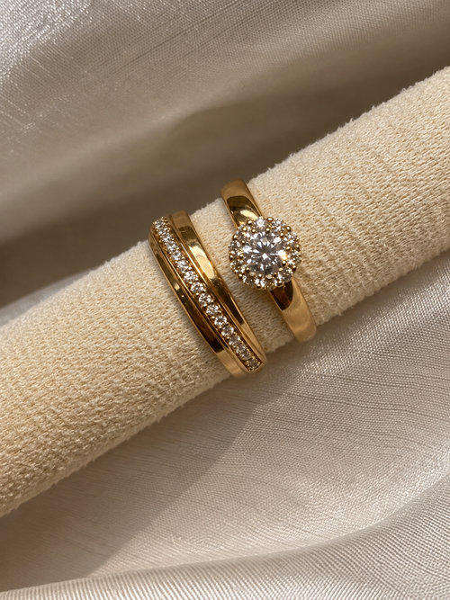 Three gold rings displayed on a textured cream fabric ring holder. One ring features a round brilliant-cut diamond in a halo setting, while the other two are slim bands, one with a pavé band and one with a curved row of small diamonds.