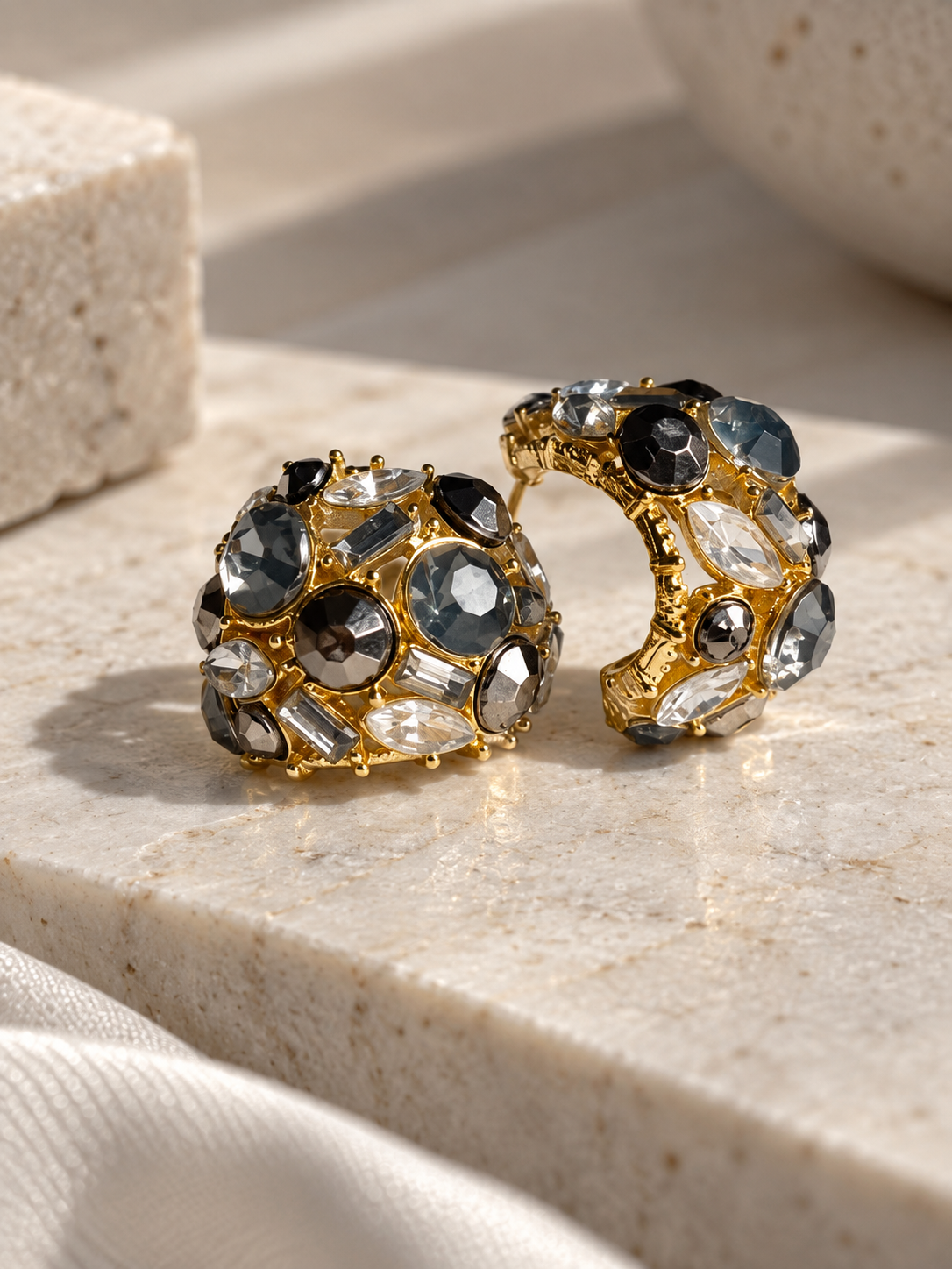 Gold-tone statement hoop earrings featuring black, smoky gray, and clear faceted crystals in a bold, intricate design, displayed on a cream marble surface.