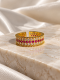 A wide gold cuff ring featuring a central row of round clear stones and a row of vivid pink gemstones, displayed on a polished cream marble slab with draped satin fabric in the background.