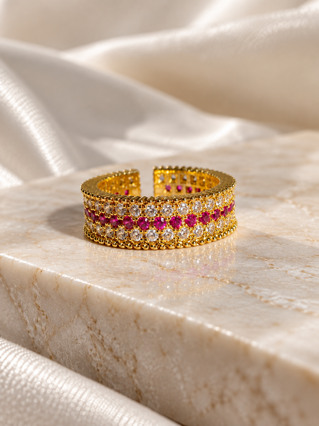 A wide gold cuff ring featuring a central row of round clear stones and a row of vivid pink gemstones, displayed on a polished cream marble slab with draped satin fabric in the background.
