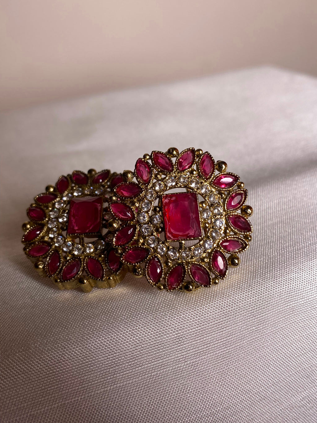 A pair of ornate gold-toned earrings with rectangular red gemstones and surrounding marquise-shaped red stones set with small clear stones, displayed on a light beige fabric surface.