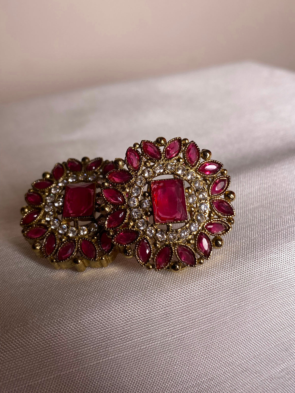 A pair of ornate gold-toned earrings with rectangular red gemstones and surrounding marquise-shaped red stones set with small clear stones, displayed on a light beige fabric surface.
