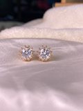 A pair of gold-toned stud earrings with clear round gemstones in a sunburst setting, resting on a soft white satin fabric.