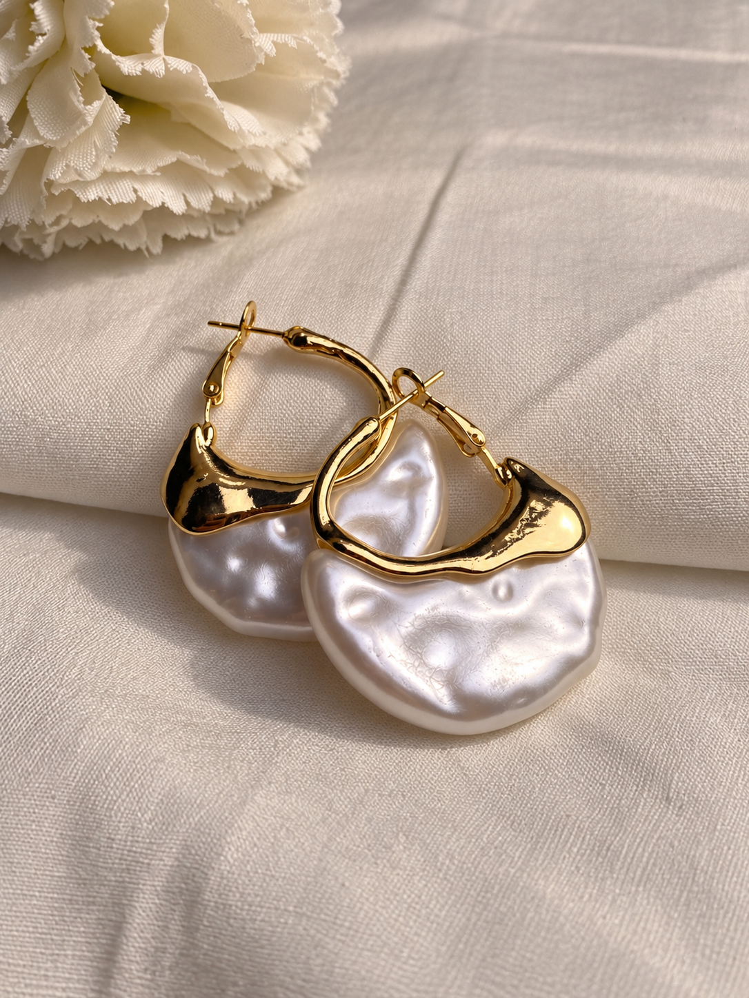 Gold-tone chunky hoop earrings with glossy white sculpted resin drops in an organic shape, displayed on soft cream fabric.