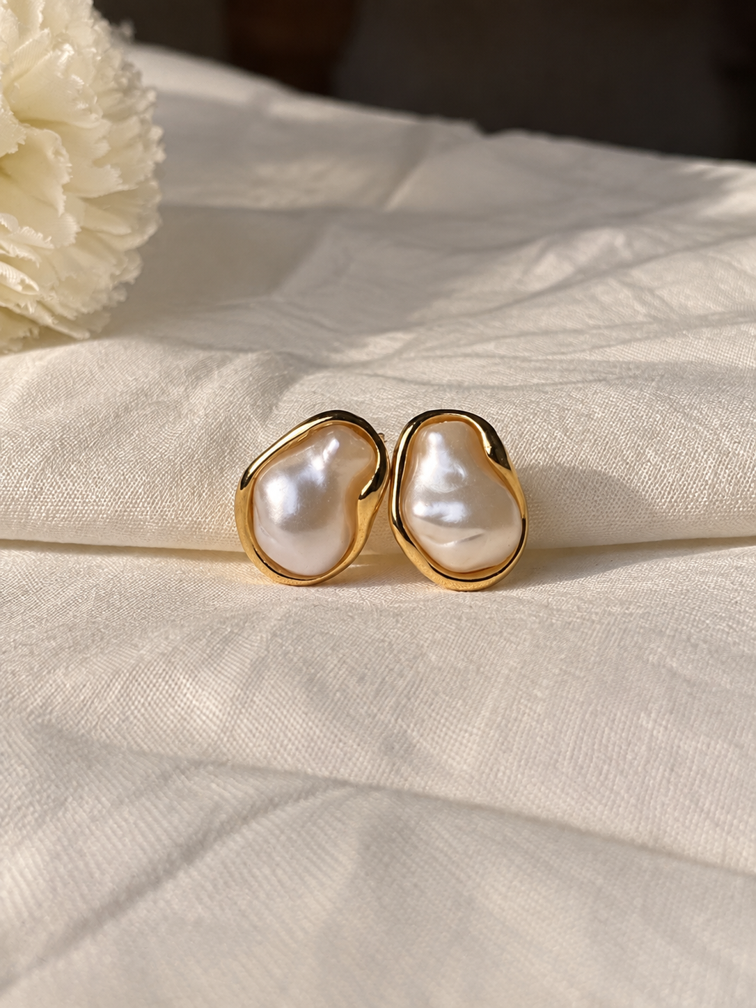 A pair of gold-toned stud earrings with irregular, organic-shaped white baroque pearls, resting on a textured cream linen fabric.