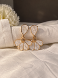 White floral drop earrings with textured petal details, gold-tone outlines, and dangling layered flower shapes on cream fabric.