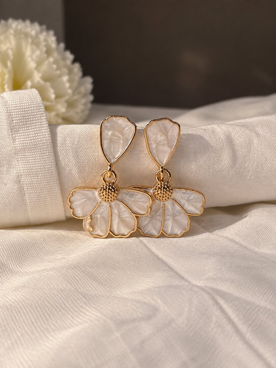 White floral drop earrings with textured petal details, gold-tone outlines, and dangling layered flower shapes on cream fabric.