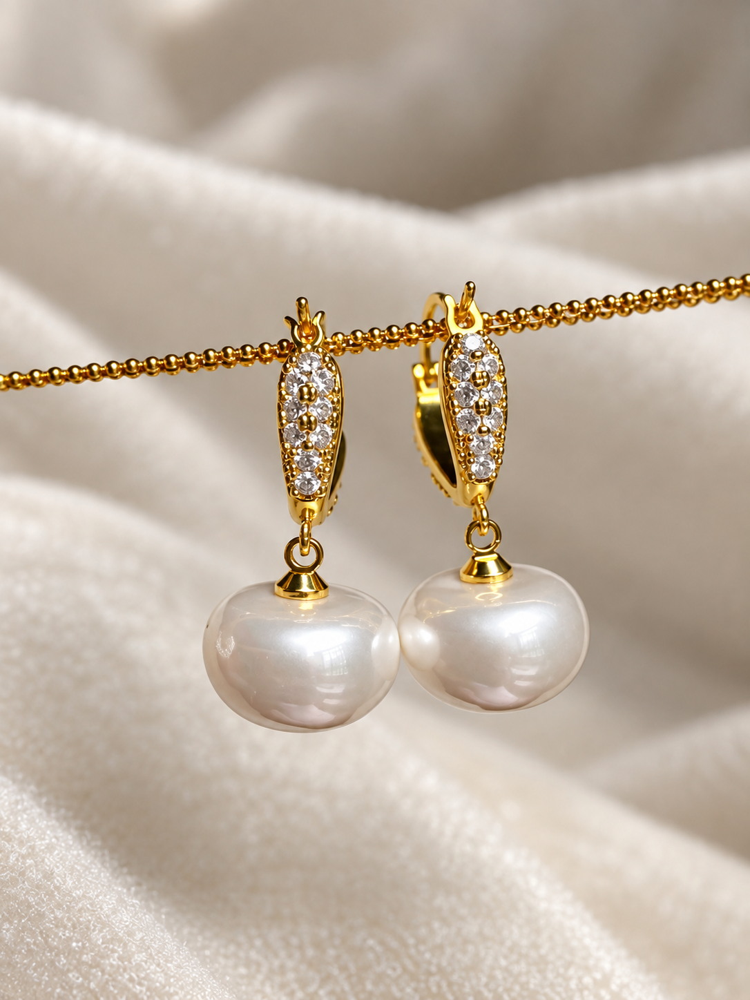 Gold-tone pearl drop earrings with sparkling clear stones and large lustrous white pearl pendants, displayed on soft cream fabric.